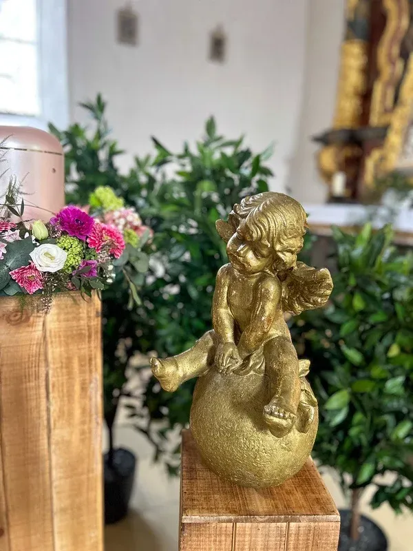 Goldene Engelsfigur auf einer Kugel neben einem Blumengesteck auf Holzsäulen in der Trauerhalle.