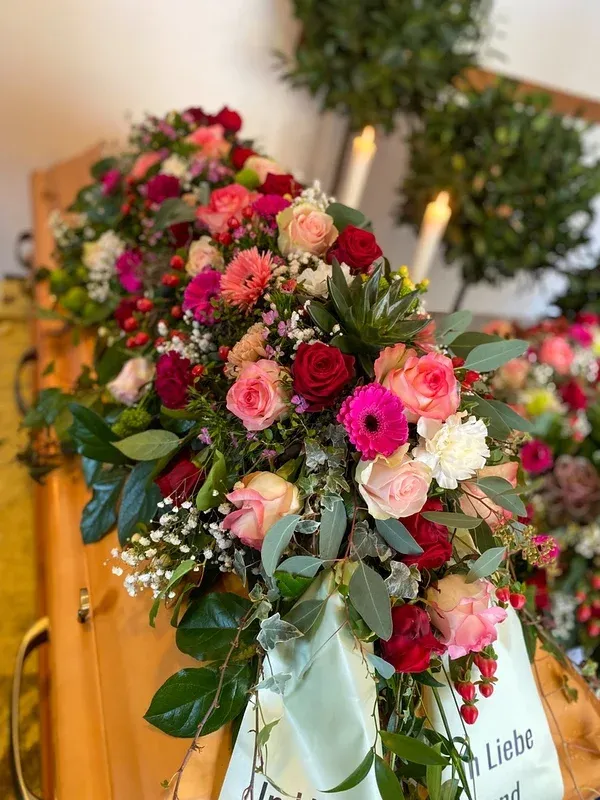Üppiges Gesteck aus rosa und roten Rosen und Gerbera auf einem Sarg im warmen Kerzenlicht.