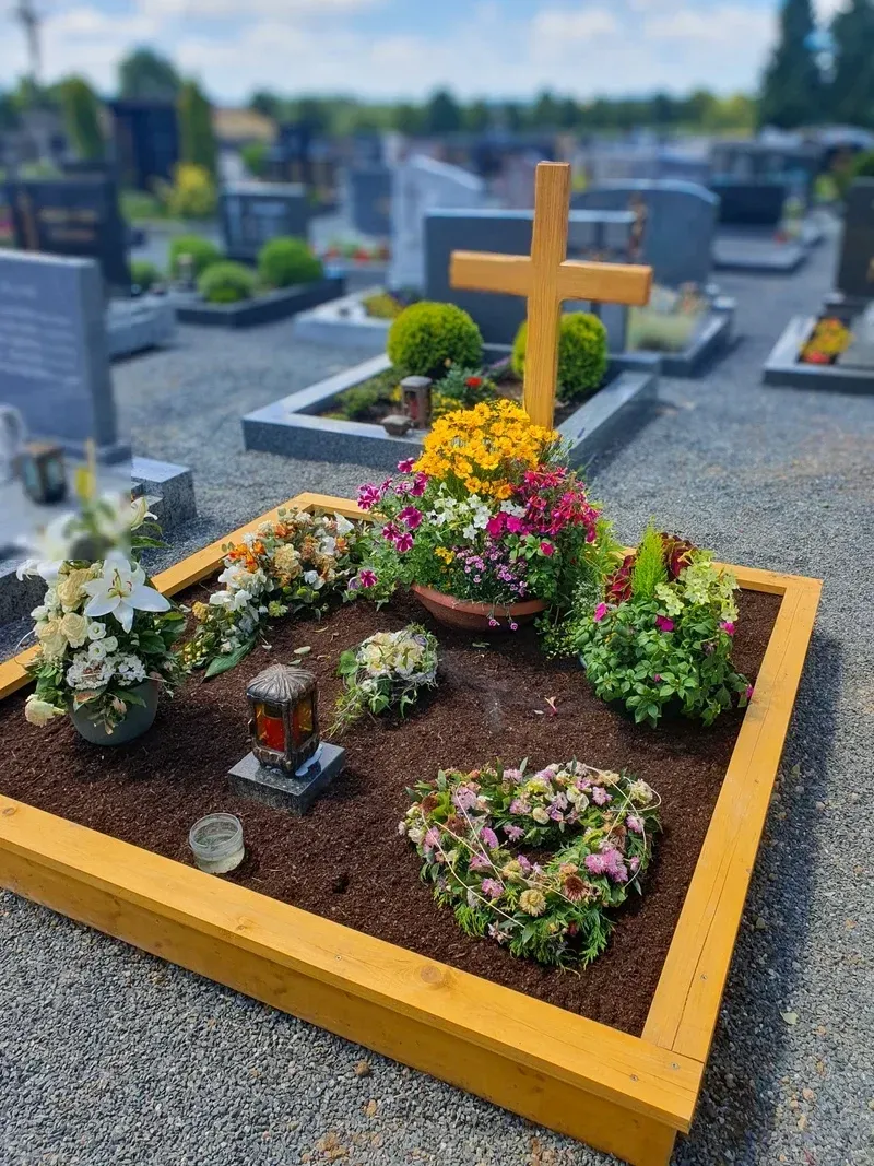 Sommerlich bepflanztes Grab mit hellem Holzkreuz, buntem Blumenschmuck und einem Herz aus Trockenblumen.