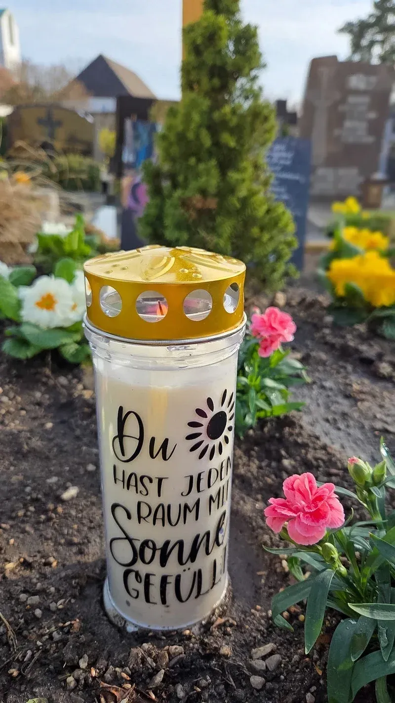 Weiße Grablicht-Kerze mit dem Spruch „Du hast jeden Raum mit Sonne gefüllt“ auf einem frühlingshaft bepflanzten Grab.