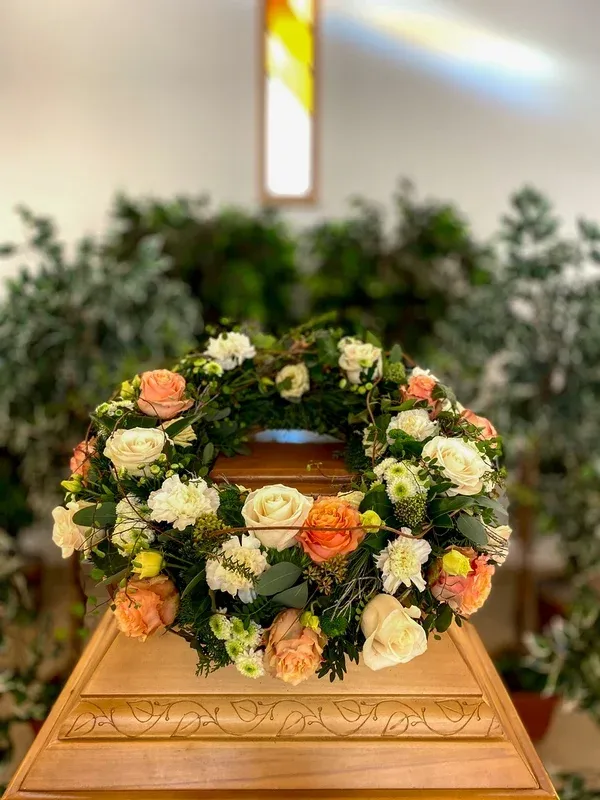 Pastellfarbener Kranz aus Rosen und Nelken auf einem Holzsarg, im Hintergrund ein leuchtendes Kirchenfenster.