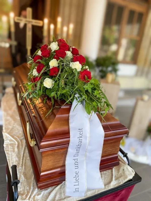 Dunkler Holzsarg mit einem Gesteck aus roten und weißen Rosen und der Schleife „In Liebe und Dankbarkeit“ vor einem Kruzifix.