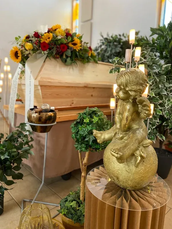 Heller Holzsarg mit Sonnenblumen-Gesteck, davor eine goldene Engelsfigur und Kerzen.