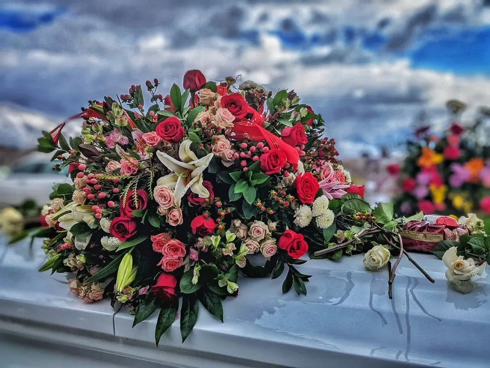 Sarggesteck aus roten und rosafarbenen Rosen, weißen Lilien und Chrysanthemen auf einem hellen Sarg.