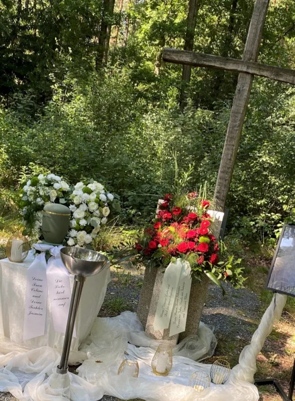 Abschiedsstelle im Wald unter einem hölzernen Kreuz: links eine dunkle Urne mit weißem Blumengesteck auf einem verhüllten Tisch, rechts ein rotes Gesteck mit Trauerschleifen auf einem Granitsockel.