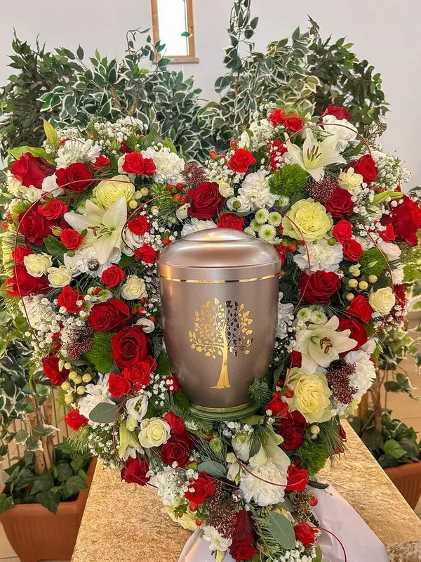 Graue Urne mit goldenem „Baum des Lebens“ in einem herzförmigen Kranz aus roten und weißen Rosen.