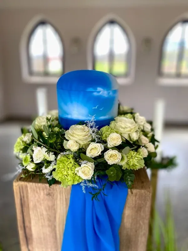 Blaue Urne mit Himmels- und Möwenmotiv, umkränzt von weißen und grünen Rosen, vor Kirchenfenstern.