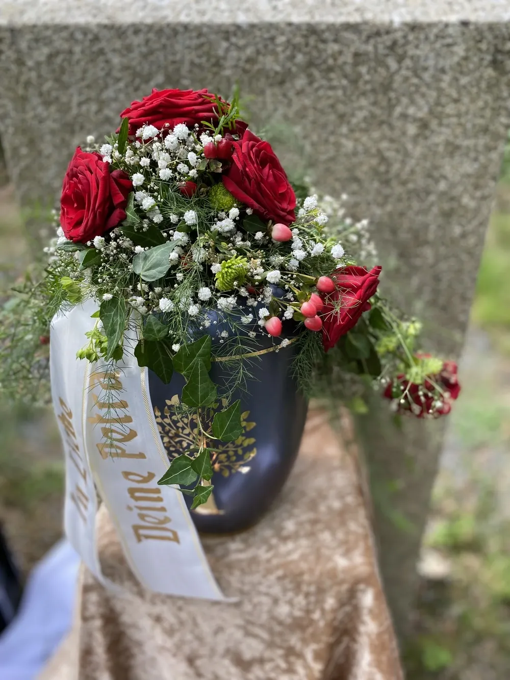 Dunkelblaue Urne mit goldenem Baummotiv, geschmückt mit roten Rosen und weißem Schleierkraut.