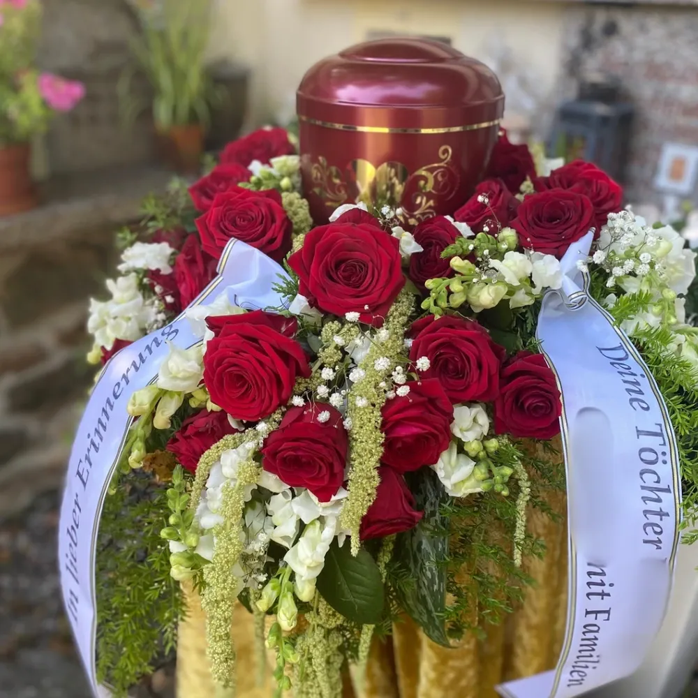 Bordeauxrote Urne mit goldenem Ornament, umgeben von einem Gesteck aus roten Rosen und weißen Blüten mit Trauerschleifen.