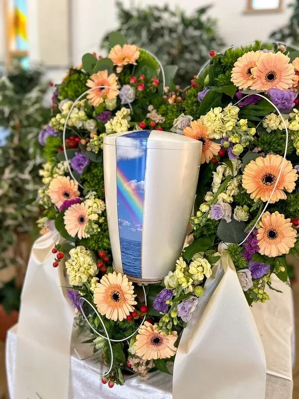 Weiße Urne mit Regenbogen- und Meeresmotiv, umkränzt von pastellfarbenen Gerbera und Rosen.
