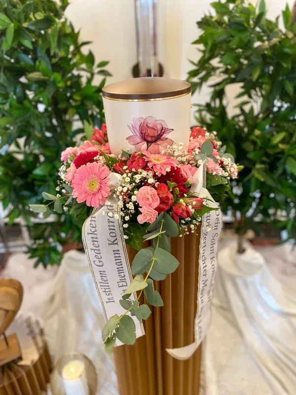 Weiße Urne mit Rosenblüten-Motiv, umgeben von einem Gesteck aus rosa Gerbera und Nelken.