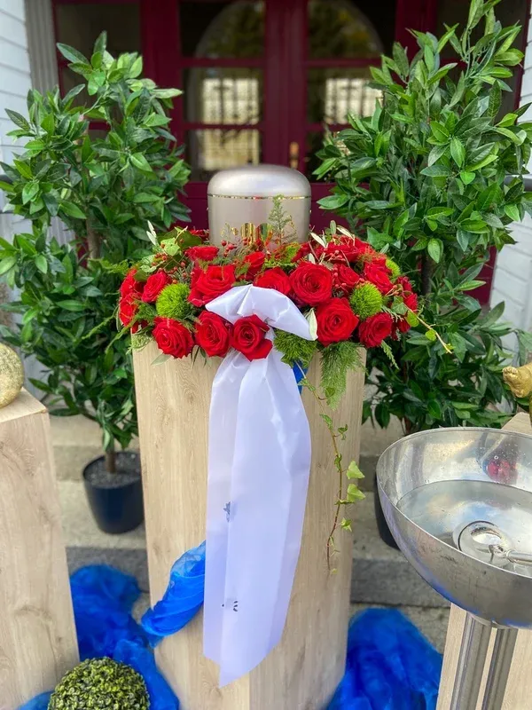 Silberne Urne mit einem Kranz aus roten Rosen auf einer Holzsäule vor einem roten Eingangsportal.