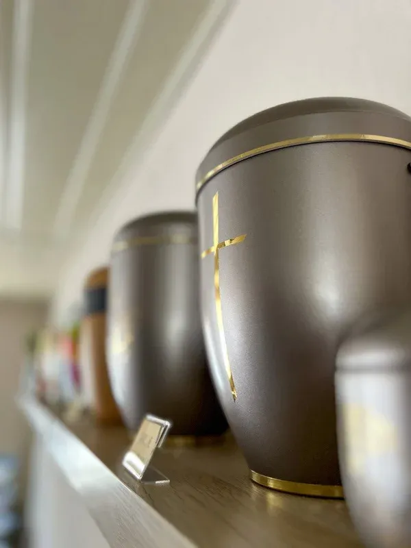 Braune Urnen mit goldenem Kreuz auf einem Ausstellungsregal in weicher Unschärfe.