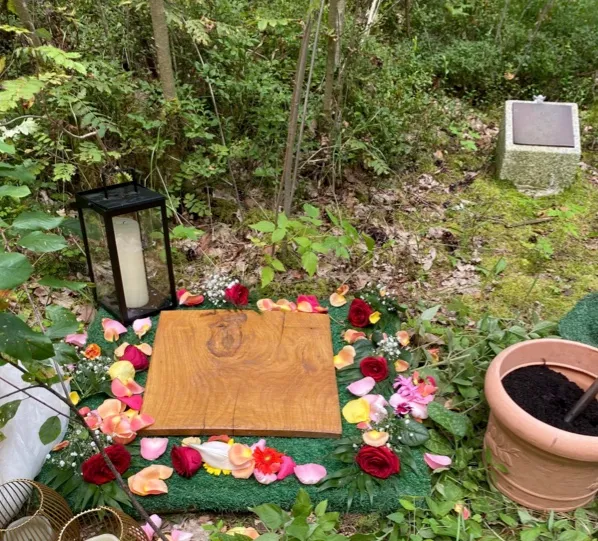 Geschlossenes Urnengrab im Wald, abgedeckt mit einer Holzplatte und umlegt von einem Kranz aus bunten Rosen- und Nelkenblüten. Daneben eine Laterne mit Kerze und ein Tontopf mit Erde.