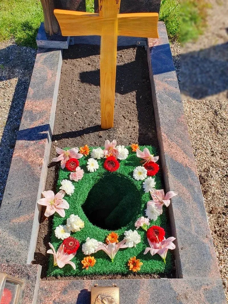 Vorbereitete Urnengrabstelle mit hellem Holzkreuz und einem Kranz aus rosa Lilien und bunten Gerbera.